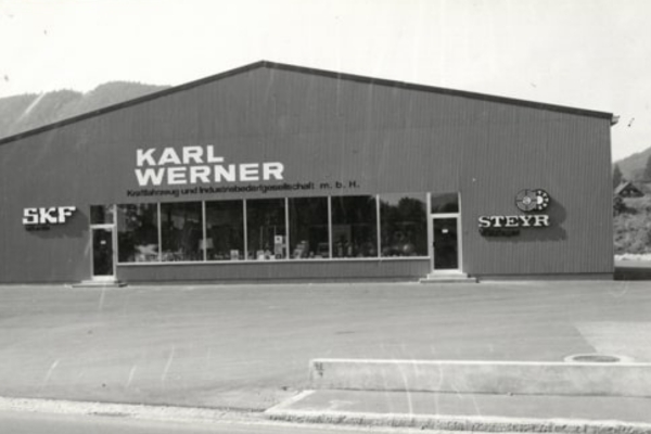 Schwarz-Weiß-Bild eines großen Industriegebäudes mit Schildern mit den Aufschriften „Karl Werner“, „SKF“ und „Steyr-Wälzlager“. Das Gebäude verfügt über markante Glasfenster, die Produkte präsentieren. Die Szene spielt an einem klaren Tag mit Bergen im Hintergrund.